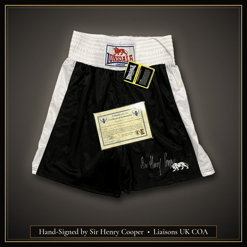 Sir Henry Cooper signed black Lonsdale London boxing shorts, size M, with swing tag and Liaisons UK Ltd COA, on a dark background with a gold border and caption