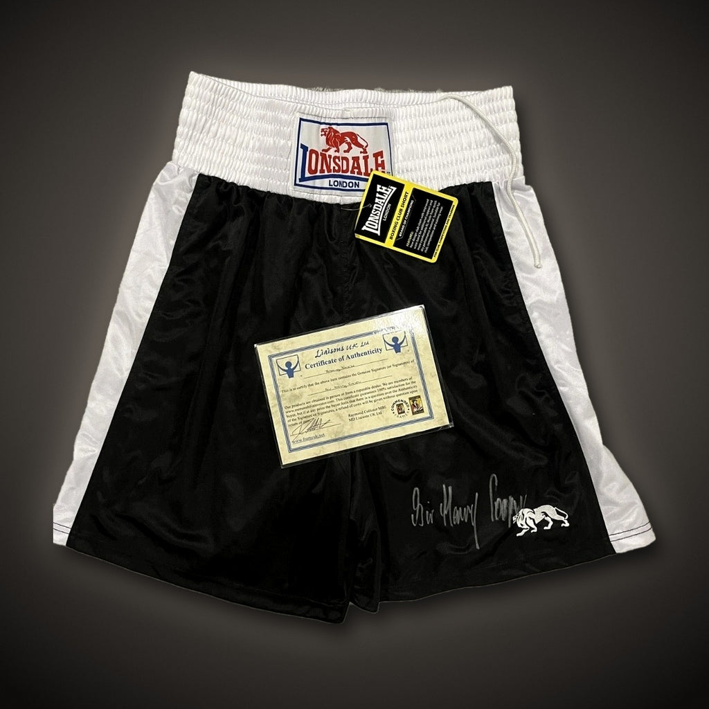 Sir Henry Cooper hand-signed Lonsdale boxing shorts, black with white side panels and white waistband, brand new with tags, on a dark background
